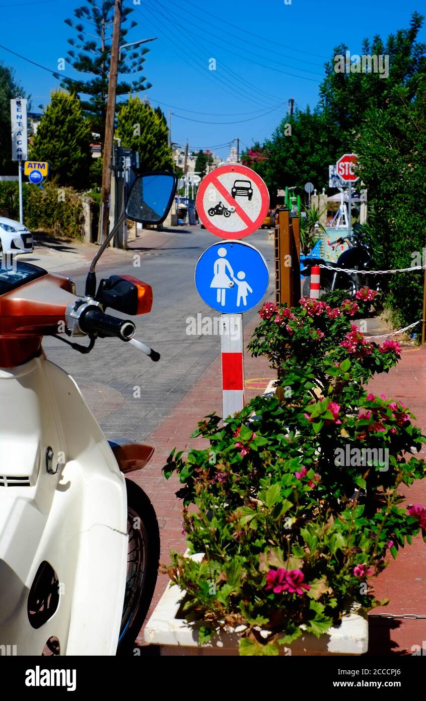 Cluttered treelined street view with scooter, road signage, plants and street furniture, Piskopiano, Crete, Greece. Stock Photo