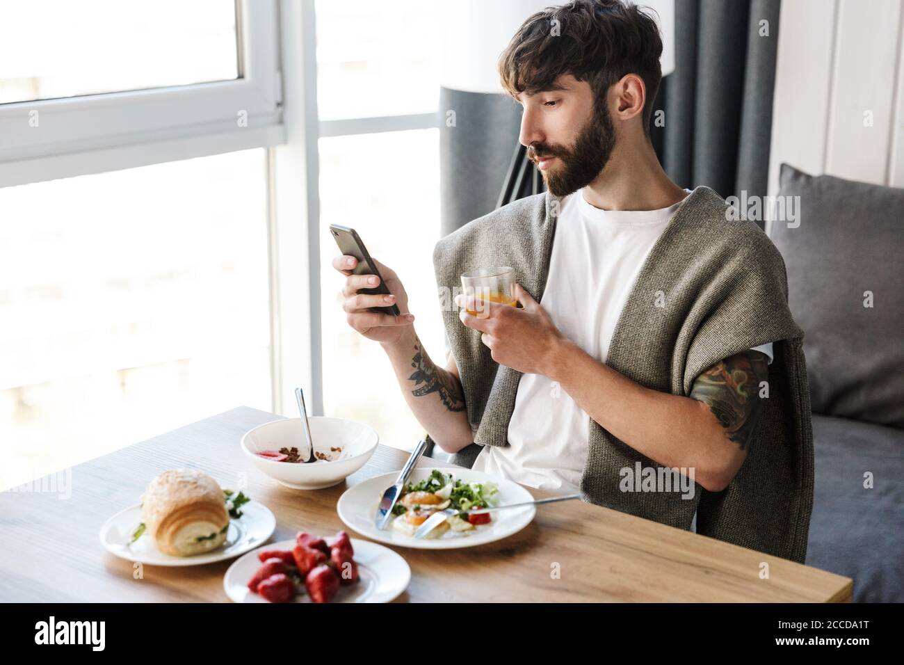 Attractive young bearded man using mobile phone while having healthy tasty dinner at home Stock Photo