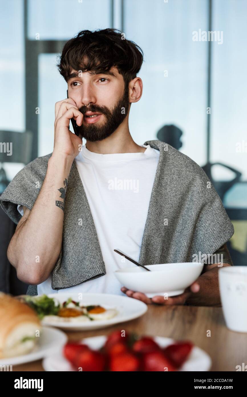 Attractive young bearded man using mobile phone while having healthy tasty dinner at home Stock Photo