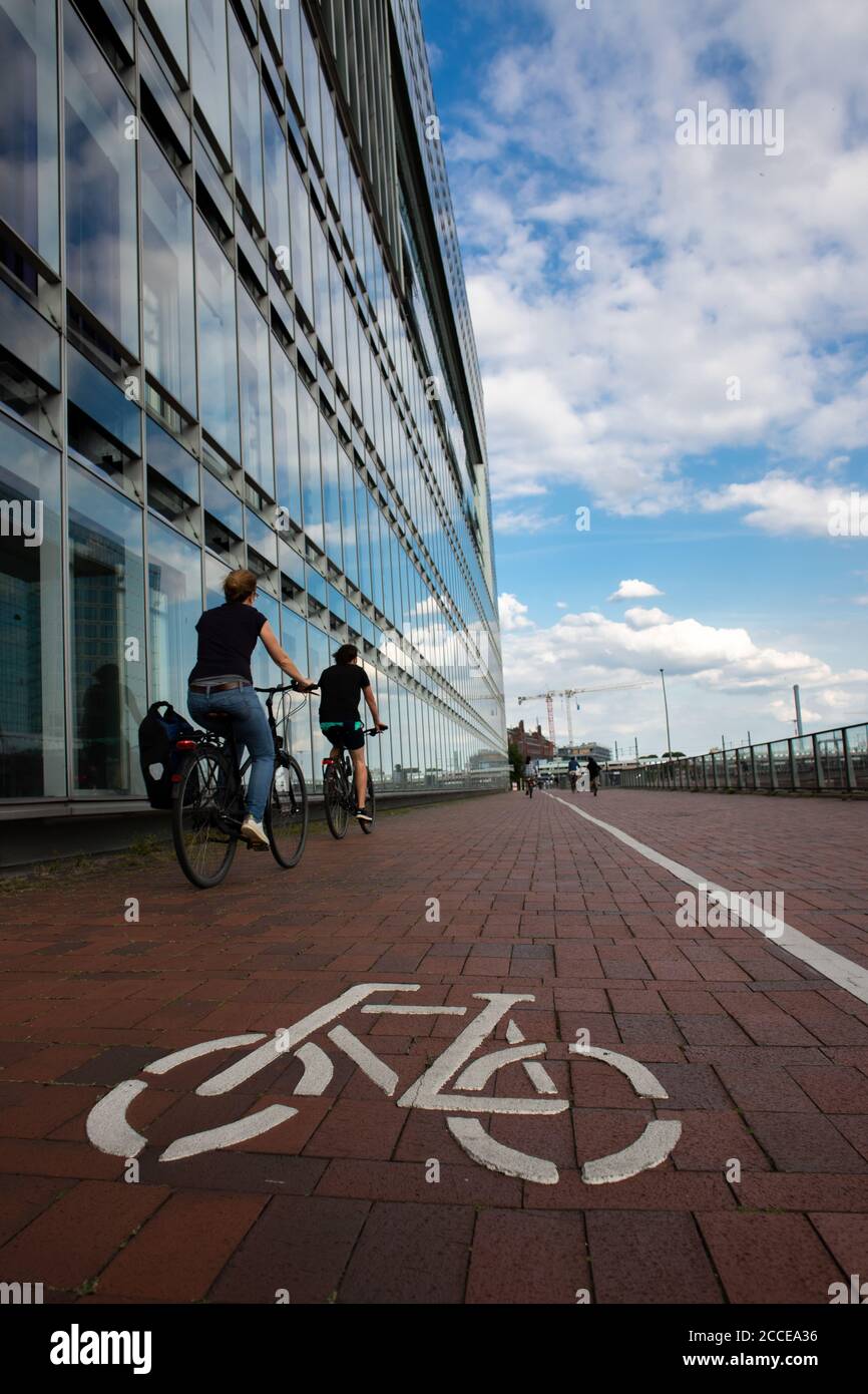 sign of a bicycle on the pavement in the foreground and modern office building, blue sky with clouds and a bicycle rider in the background, low angle, Stock Photo