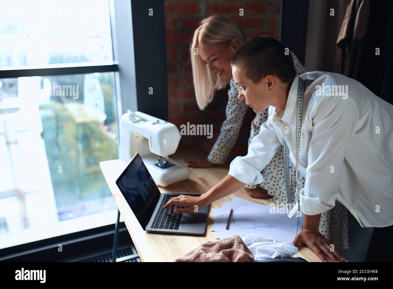 Garments Computer Work model of new dress on laptop. Two women in white clothing work on new ...