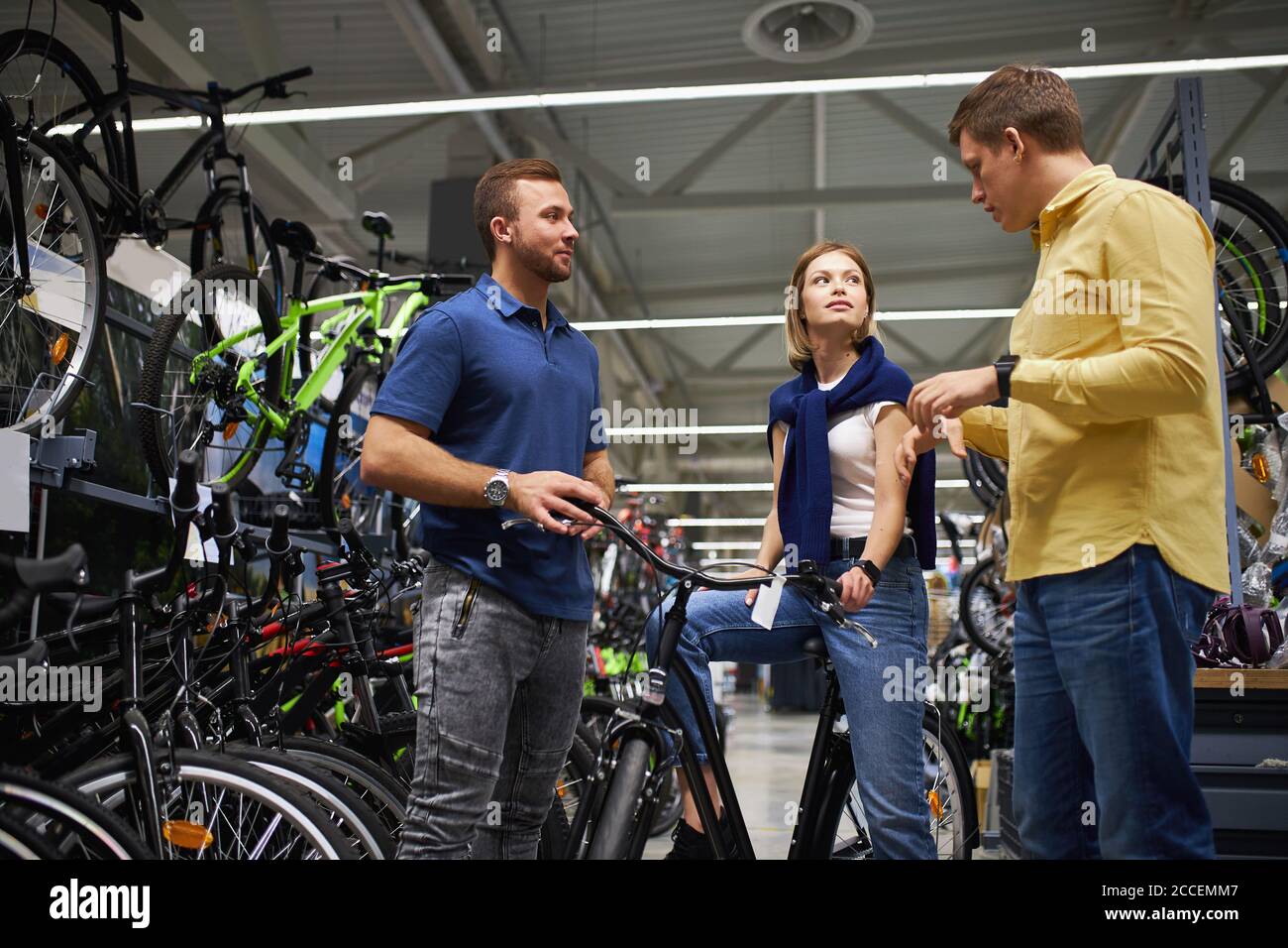 man and consultant discuss products in assortment, woman sit on sportive bicycle, she doubts, look at another Stock Photo