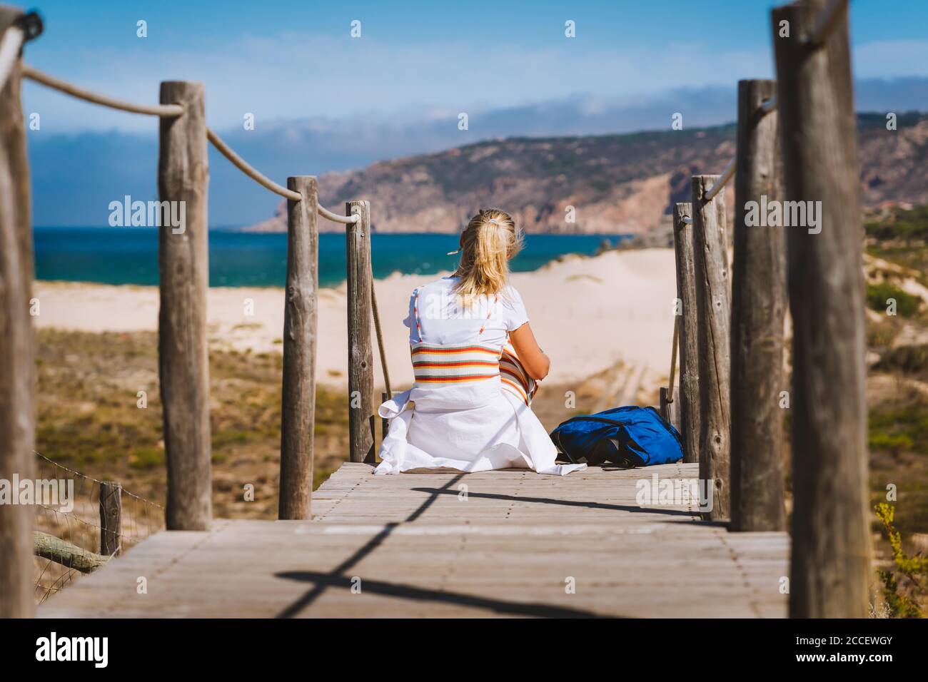 Adult female tourist sitting on path leading to Praia do Guincho Beach. Cascais, Portugal. This is popular blue flag Atlantic beach for surfing, winds Stock Photo