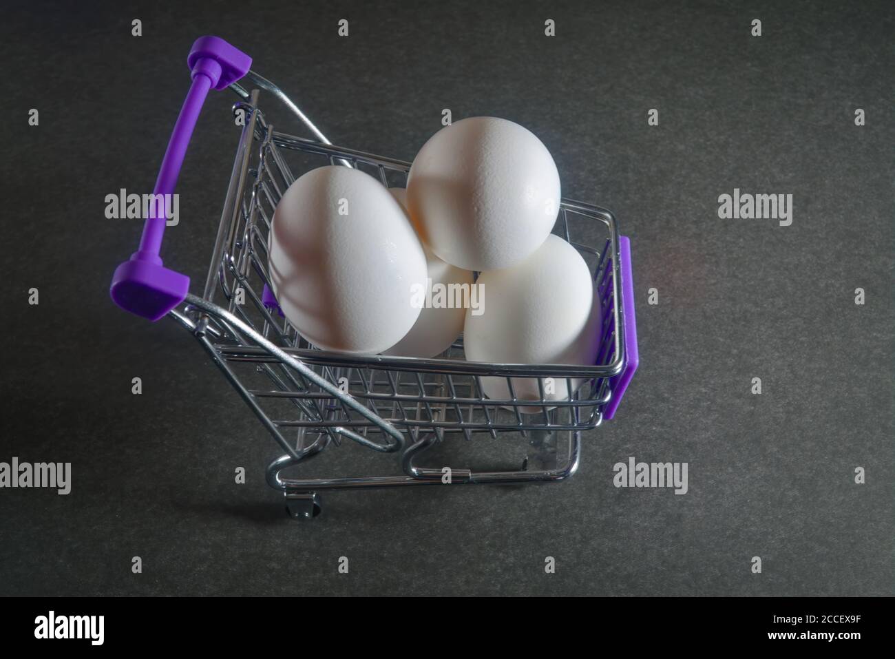 A grocery cart with three white chicken eggs on a dark gray background close-up. Stock Photo