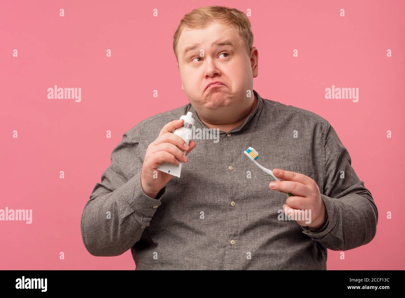 Questioning doubtful man with curved lips looking aside while holding toothbrush for cleaning teeth in hands, posing isolated over pink background. Stock Photo