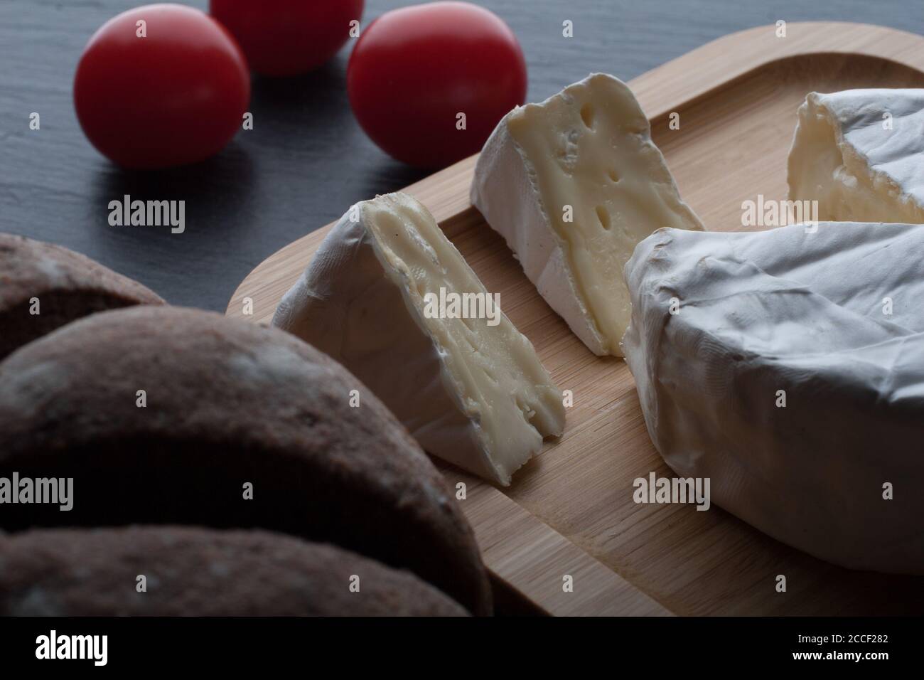 Fresh French Brie cheese and a one slice on a bamboo board with cherry tomatoes, bread on the black plate. Stock Photo