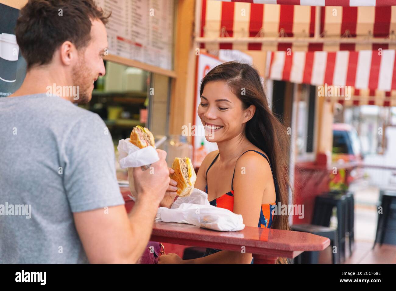 Happy couple eating sandwiches in typical retro cafe in Florida. Cuba sandwich local food. Summer travel tourist lifestyle young Asian woman smiling Stock Photo