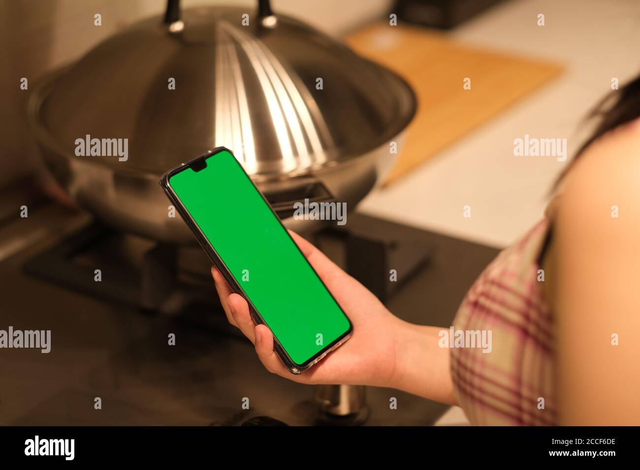 over shoulder view of woman holding green screen phone in kitchen. Stock Photo