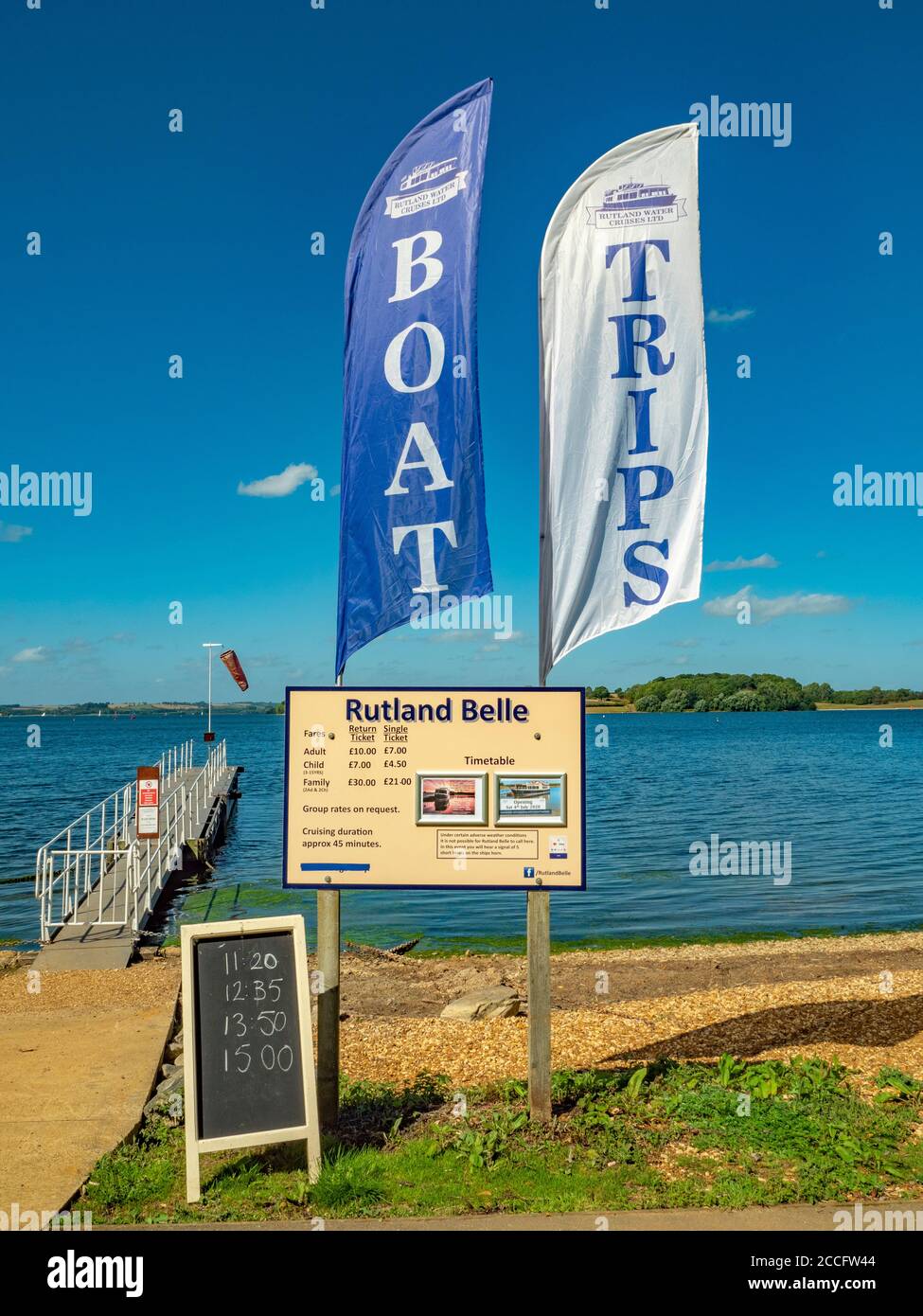 Tour boat trips on Rutland Water, with notice board of cost and timetable, and a pair of sails flying next to the passenger jetty, against a blue sky. Stock Photo