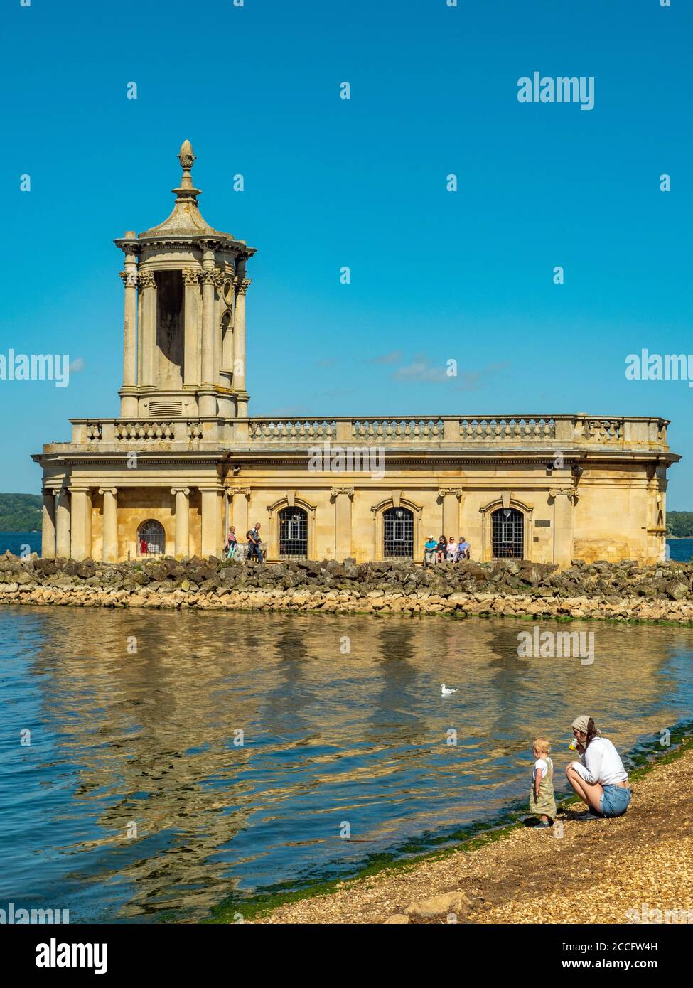Historic Normanton Church standing at the end of a causeway on Rutland Water, with visitors outside in the sunshine against a blue sky. England, UK. Stock Photo