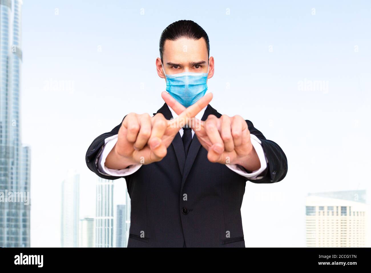 Prohibit & forbid. Businessman agent doing a ban hand gesture at the camera wearing a medical face mask and suit with the city in the background. Stock Photo