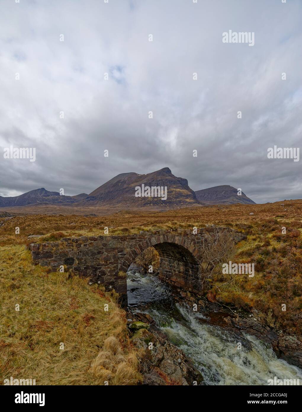 Fast flowing water coming through an arch of the old Stone road Bridge, with Mountains in the Background on a wet day in April in the Highlands. Stock Photo