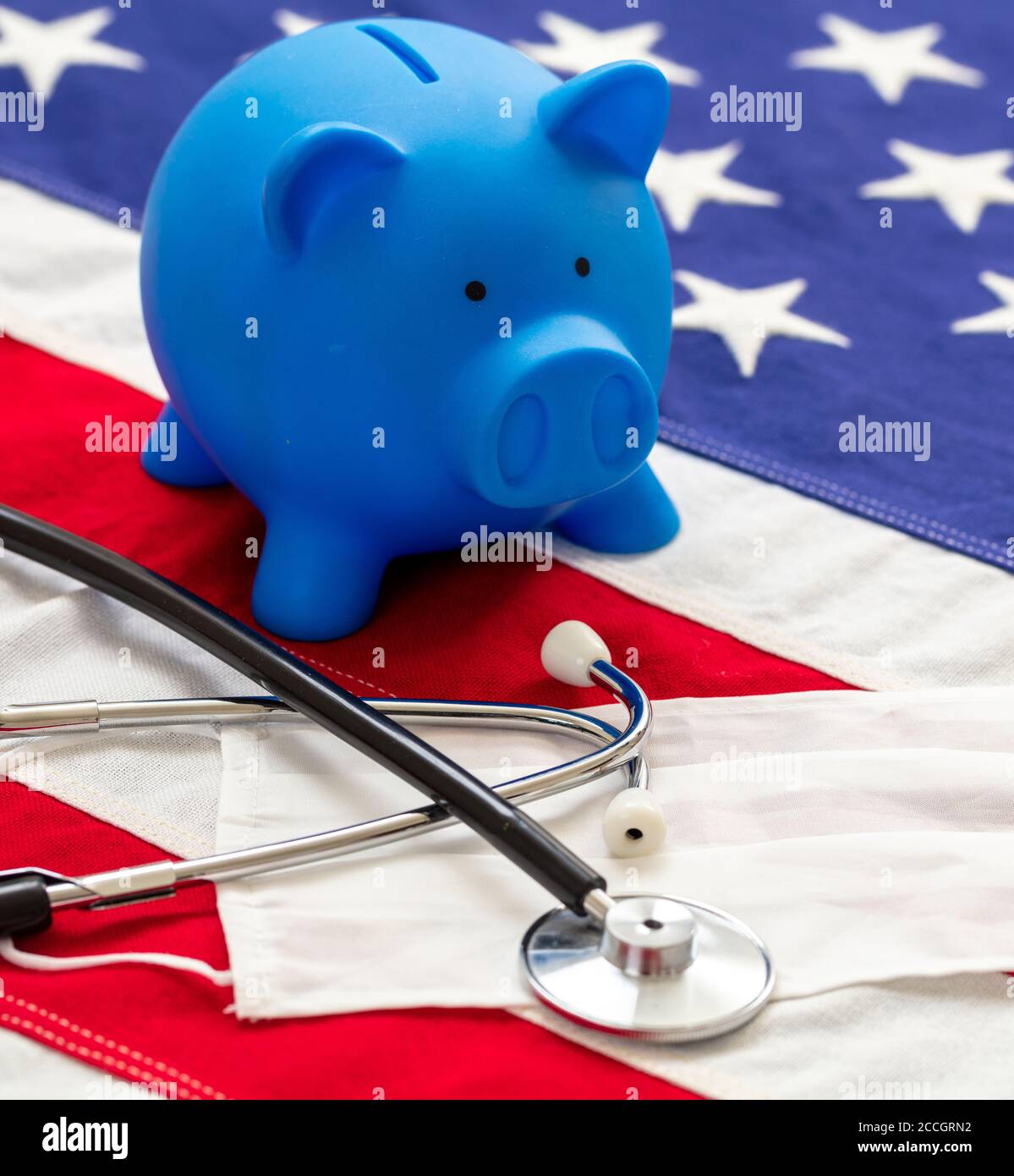 USA health care cost, coronavirus days. Medical stethoscope, protective face mask and piggy bank on a US of America flag, banner. Stock Photo