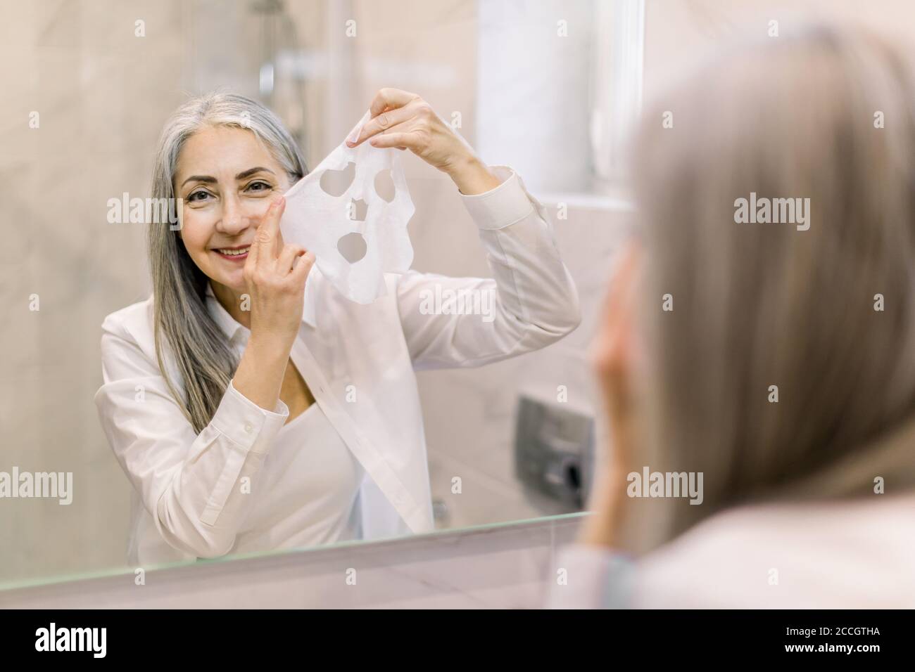 Smiling senior attractive woman is applying sheet mask on her face in the bathroom. Fabric cloth face mask to improve skin condition with antiage Stock Photo