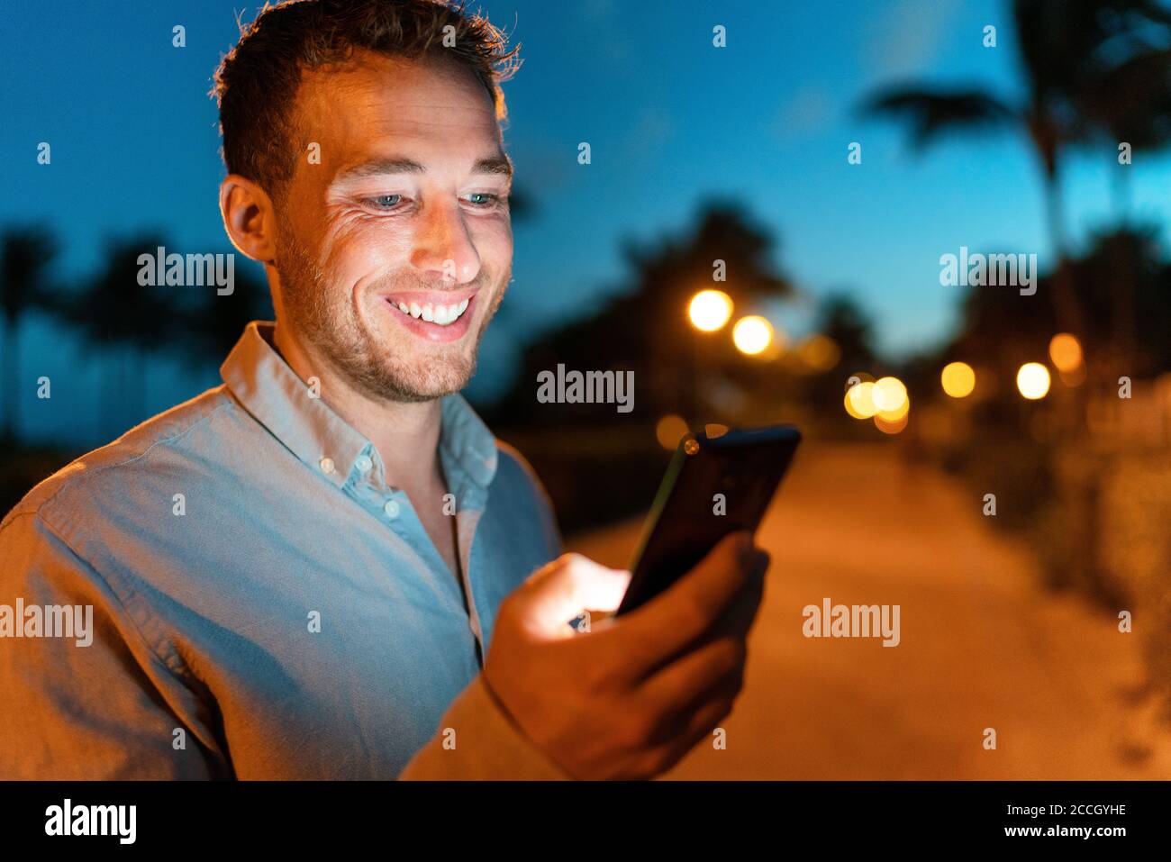 Man smiling looking at phone outside on city street at night texing online using smartphone. Young male face lit by screen light using mobile Stock Photo