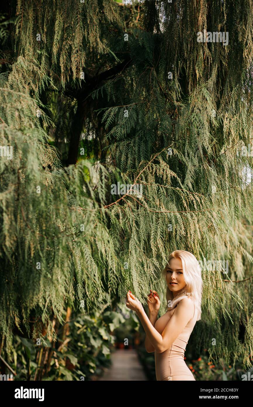 Beautiful smiling slender woman in flesh color swimsuit standing under leaves of big exotic tree. Slim blonde lady happy to spending vacation at resor Stock Photo