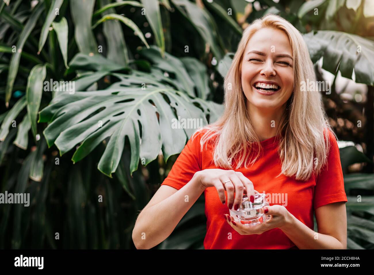 Beautiful delighted caucasian blonde woman with healthy smooth clean skin holding jar of cream, standing on tropical forest background. Concept of nat Stock Photo