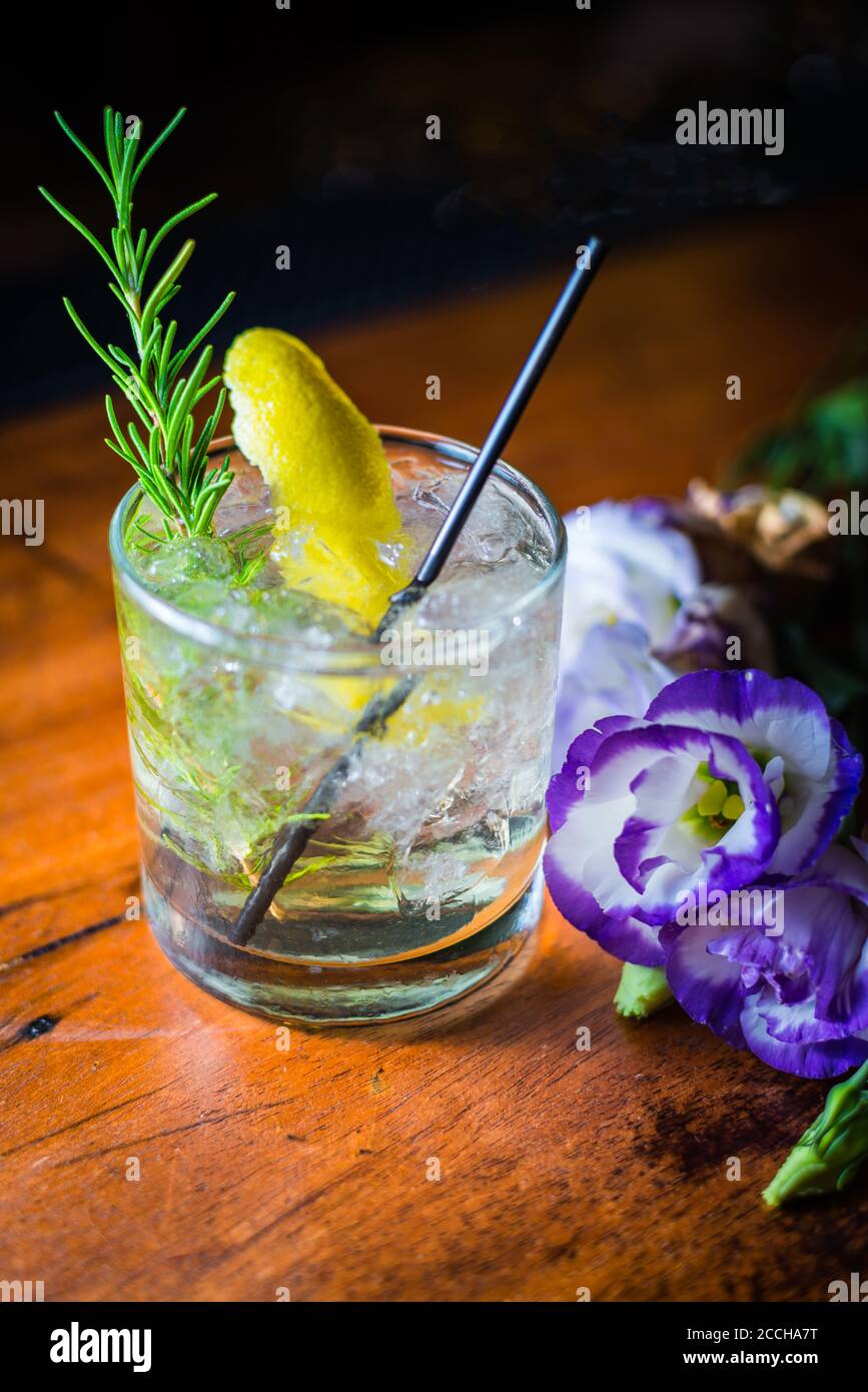 An elegant looking craft cocktail in rocks glass with lemon & rosemary garnish sits next to a flower in a sophisticated, classy, rustic bar setting. Stock Photo