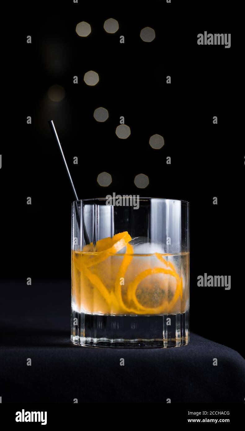 Classy, modern, sophisticated scene of a craft cocktail with orange garnish in rocks glass isolated on black background with grey bokeh circles. Stock Photo