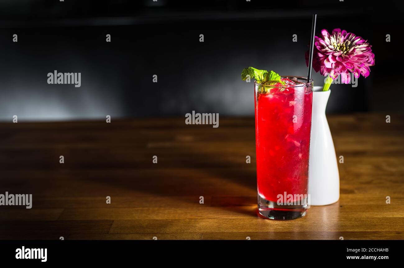 A classy, romantic scene of a red craft cocktail in a collins glass next to a flower in a white vase on a wooden table Stock Photo