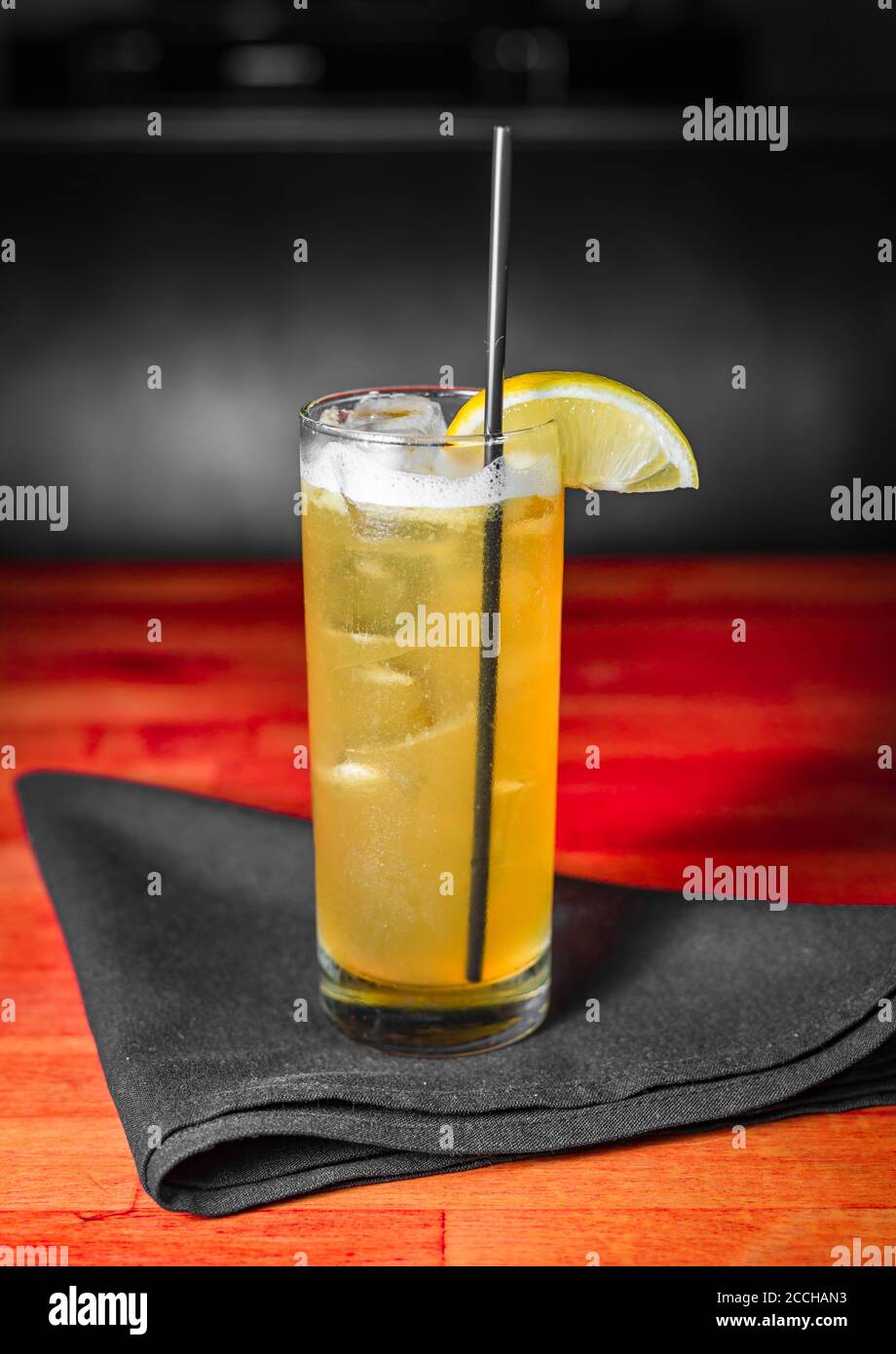 A yellow craft cocktail in a collins glass sitting on a black napkin atop a wooden table stained red Stock Photo