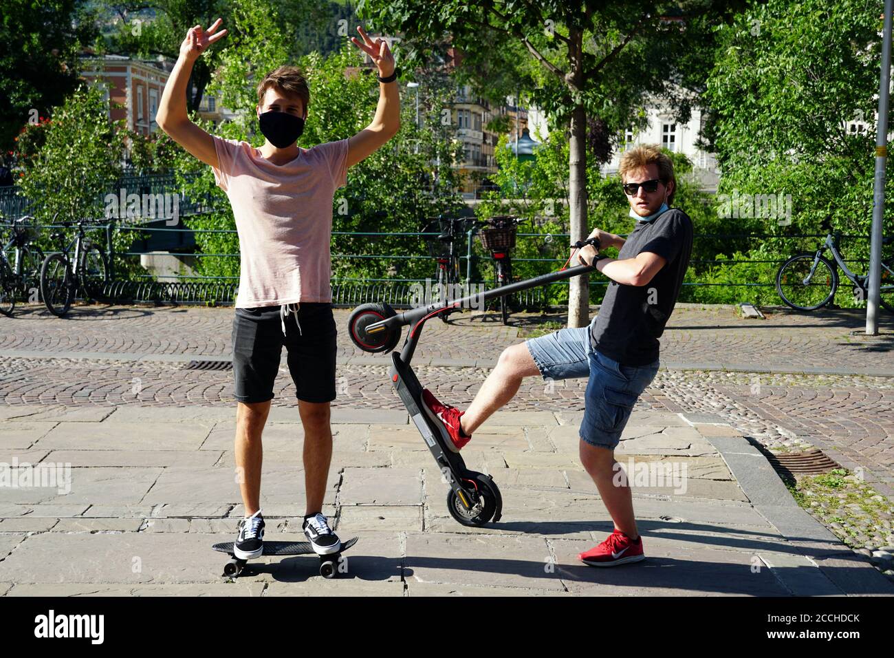 Two sporty young guys enjoying outside activities after lock down because COVID-19. One on a scateboard, the other one on an e-scooter in Merano, Sout Stock Photo