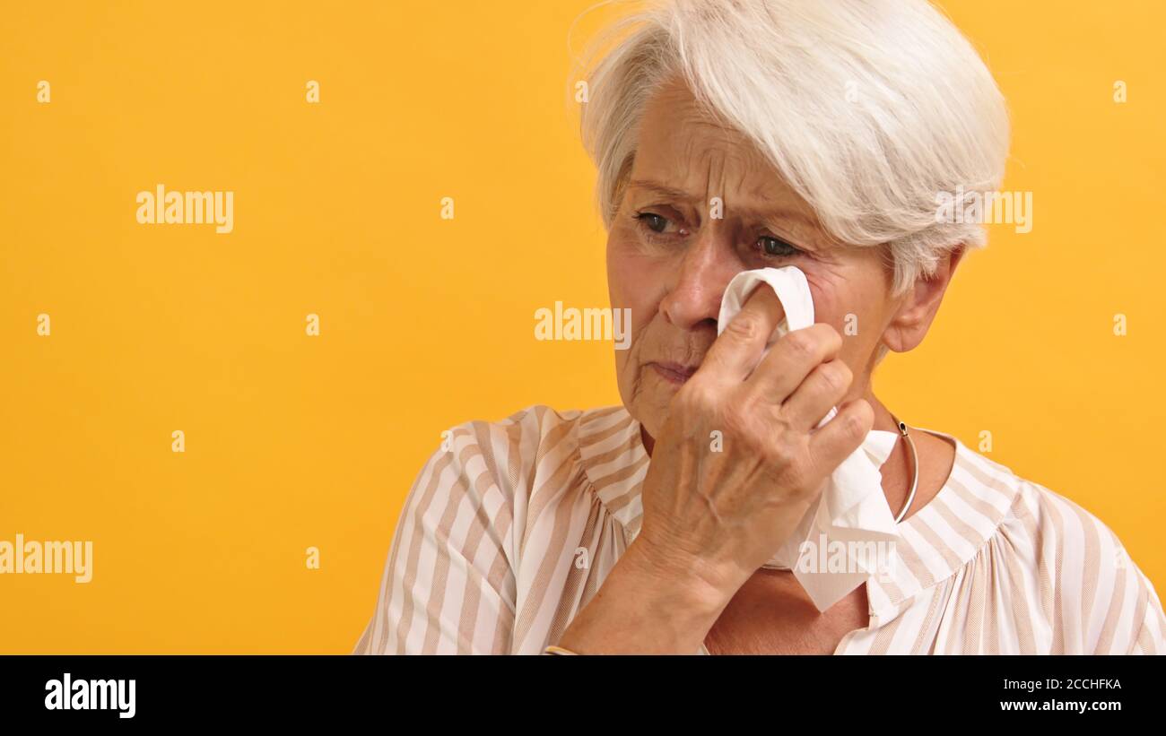 Portrait of sad vulnerable old woman wiping her tears. Isolated on the orange background. High quality photo Stock Photo