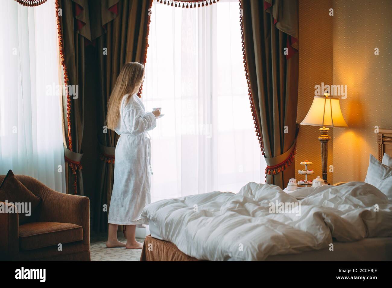 The girl Stit near the window and drinks morning coffee in the hotel room or at home. Stock Photo