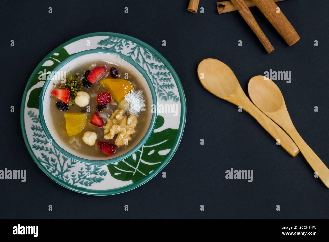 Traditional Turkish Dessert Noah's Puding in stylish green bowl on black surface with spoon and copy space.Top view. Stock Photo