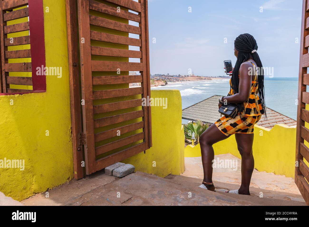 African woman dressed in colorful clothes trying to make contact with her mobile phone on a hill overlooking the coast of Accra in Ghana Stock Photo