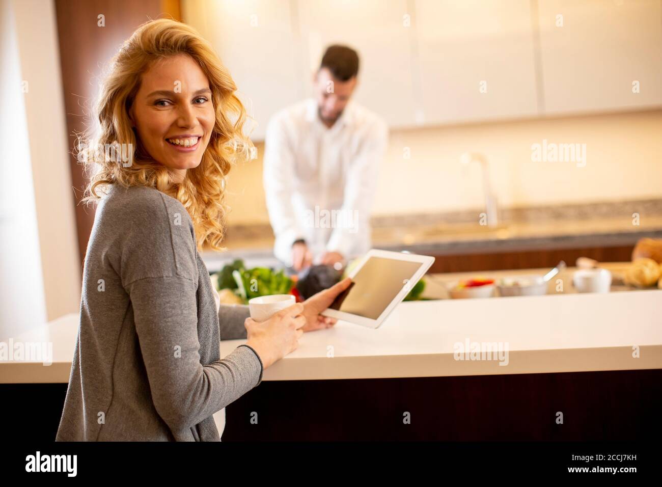 Young woman using tablet at the kitchen table while man preparing food Stock Photo