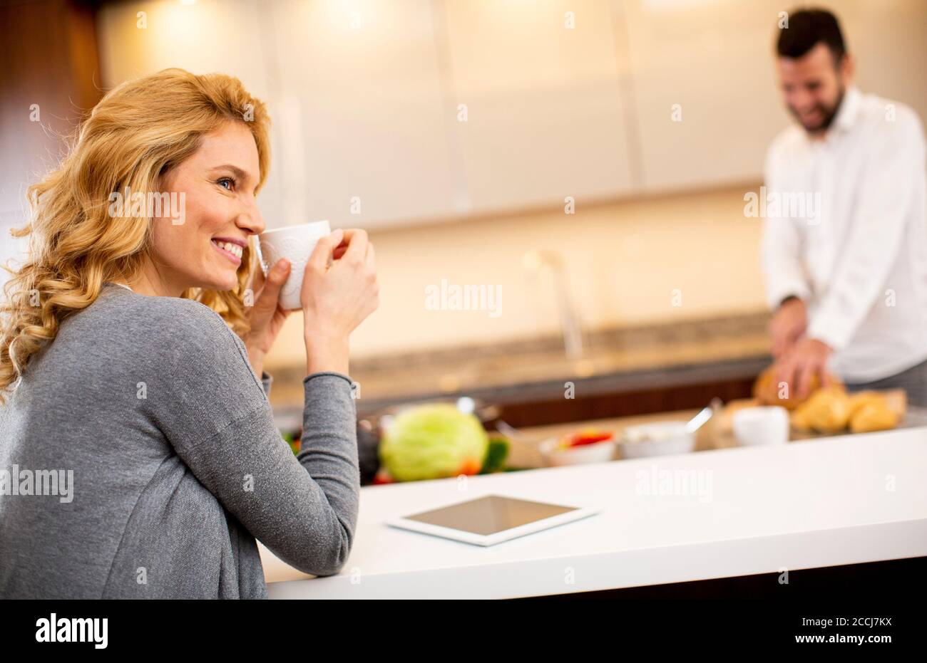 Young woman drinking coffee at the kitchen table while man preparing food Stock Photo