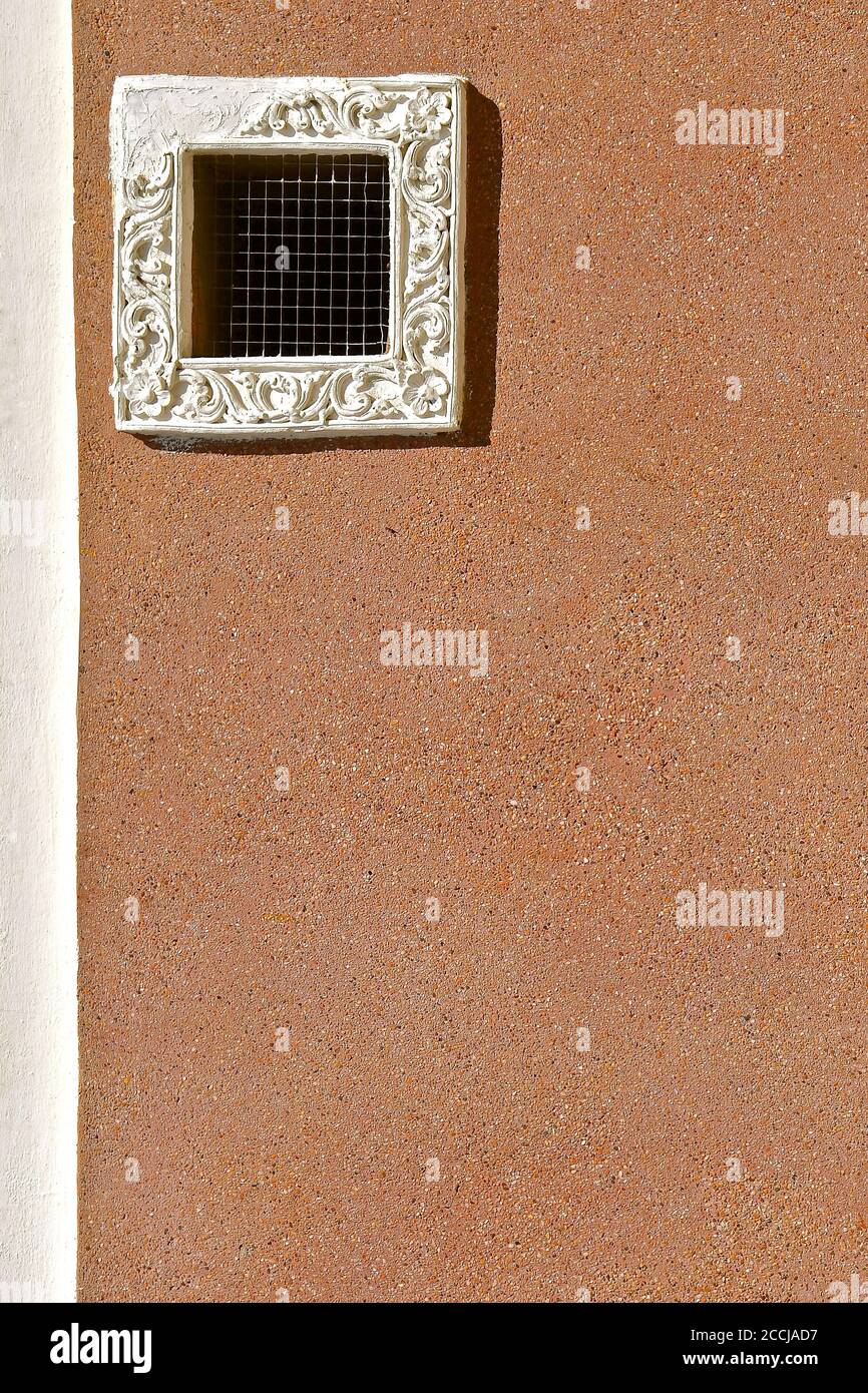 Small square ventilation vent with chicken wire grating and an ornate white plaster window frame. Isolated on a sandy coloured pebble dashed wall. Stock Photo