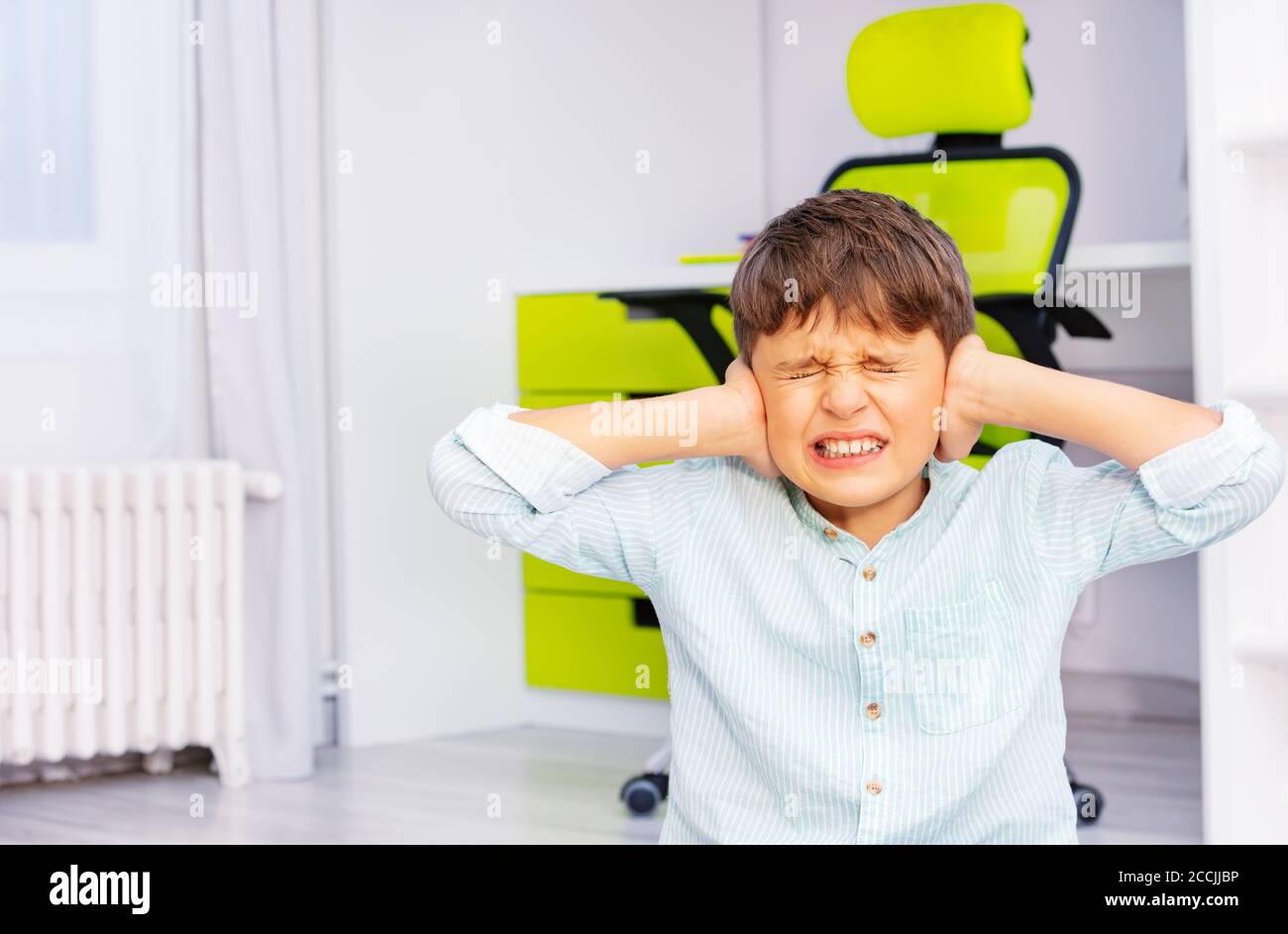 Autistic school age boy with strong negative face expression closing ears and grins teeth sitting in his room Stock Photo
