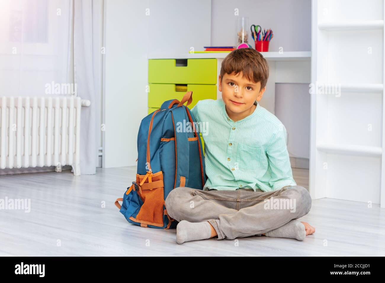 Smiling calm little boy sit in his room with positive expression holding school rucksack Stock Photo