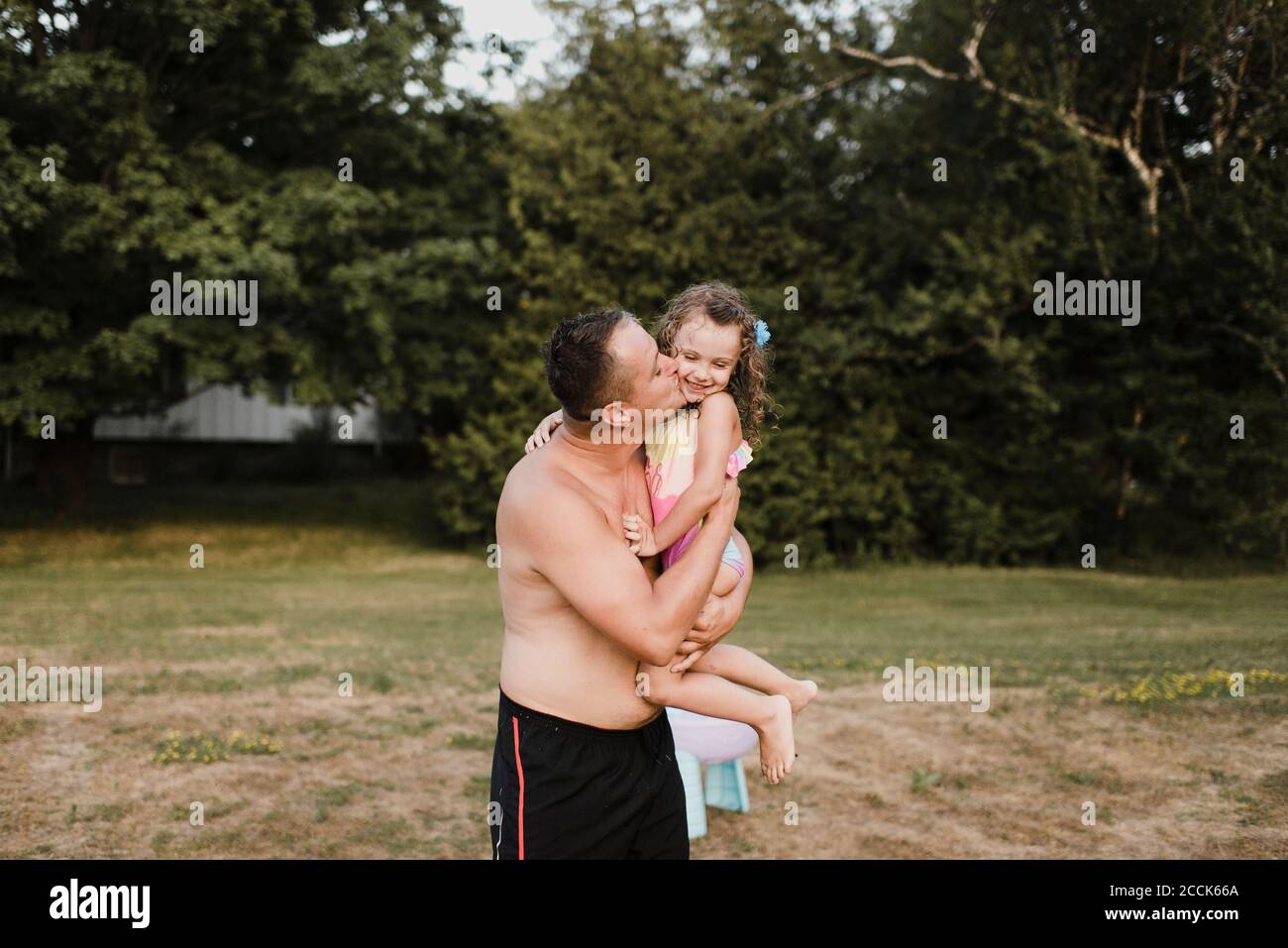 Father wearing swimming trunks carrying and kissing little daughter on a meadow Stock Photo