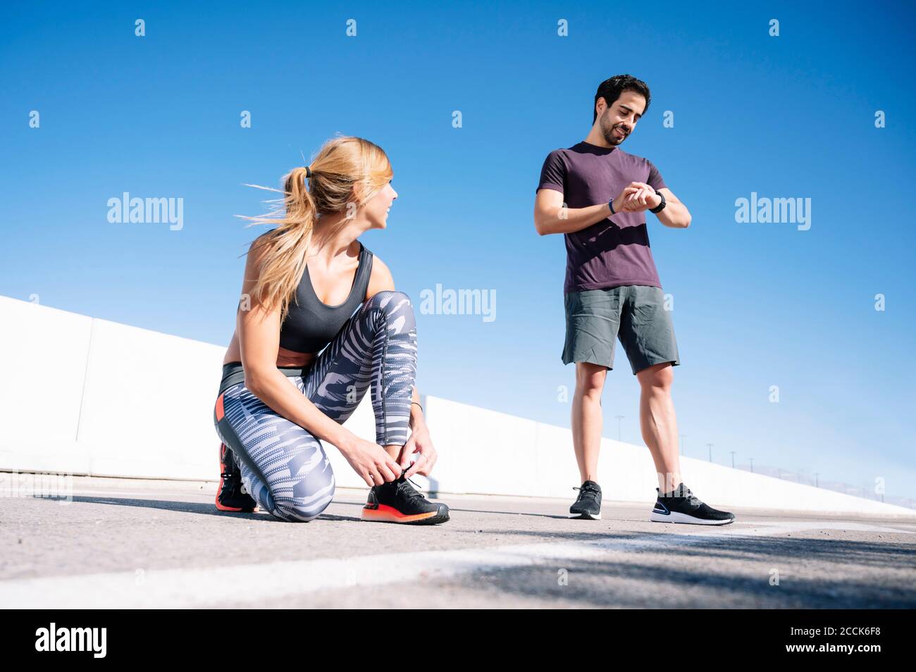 man checking time while woman tying shoelace on road against clear blue sky in city Stock Photo