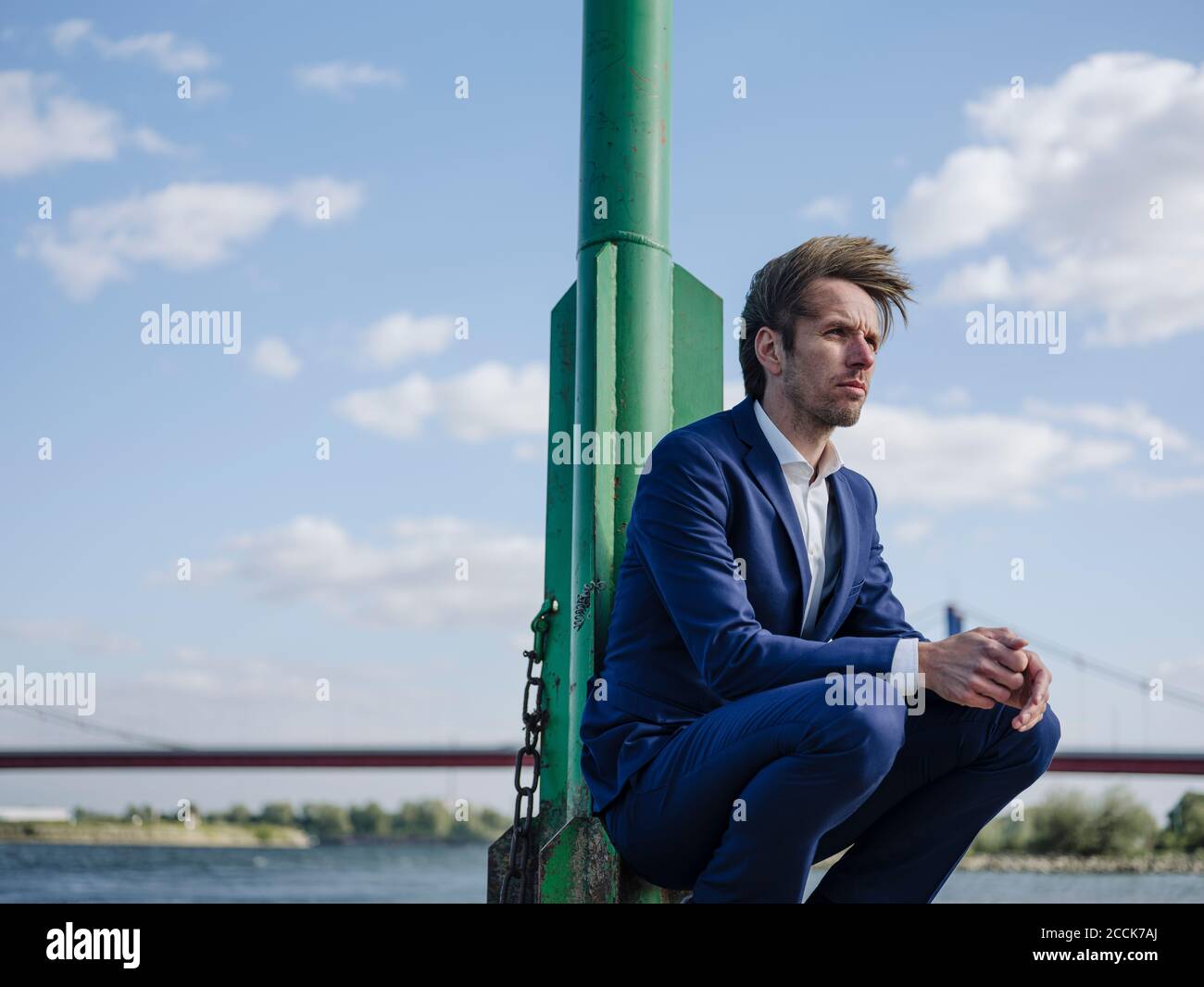 Thoughtful businessman with hands clasped crouching by pole against sky Stock Photo