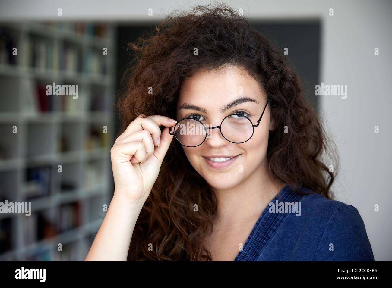 Smiling woman with long brown curly hair wearing eyeglasses at home Stock Photo