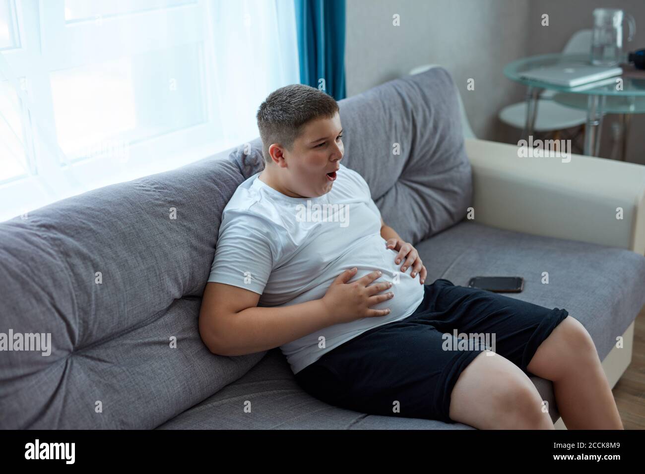 fat teenager boy has stomach ache after junk food, sit on sofa holding