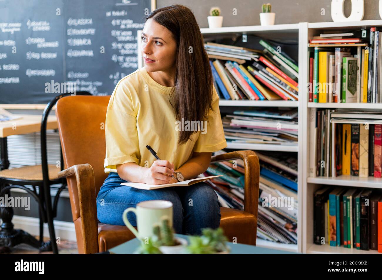 Thoughtful woman writing in book while sitting on chair in coffee shop Stock Photo