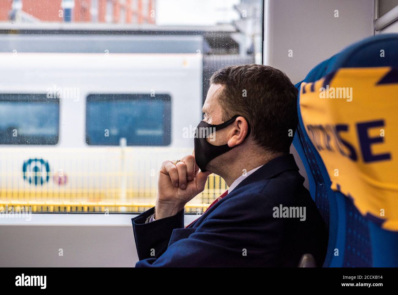A man wearing a suit and black face covering looks out of a static train window in Leeds station. Stock Photo