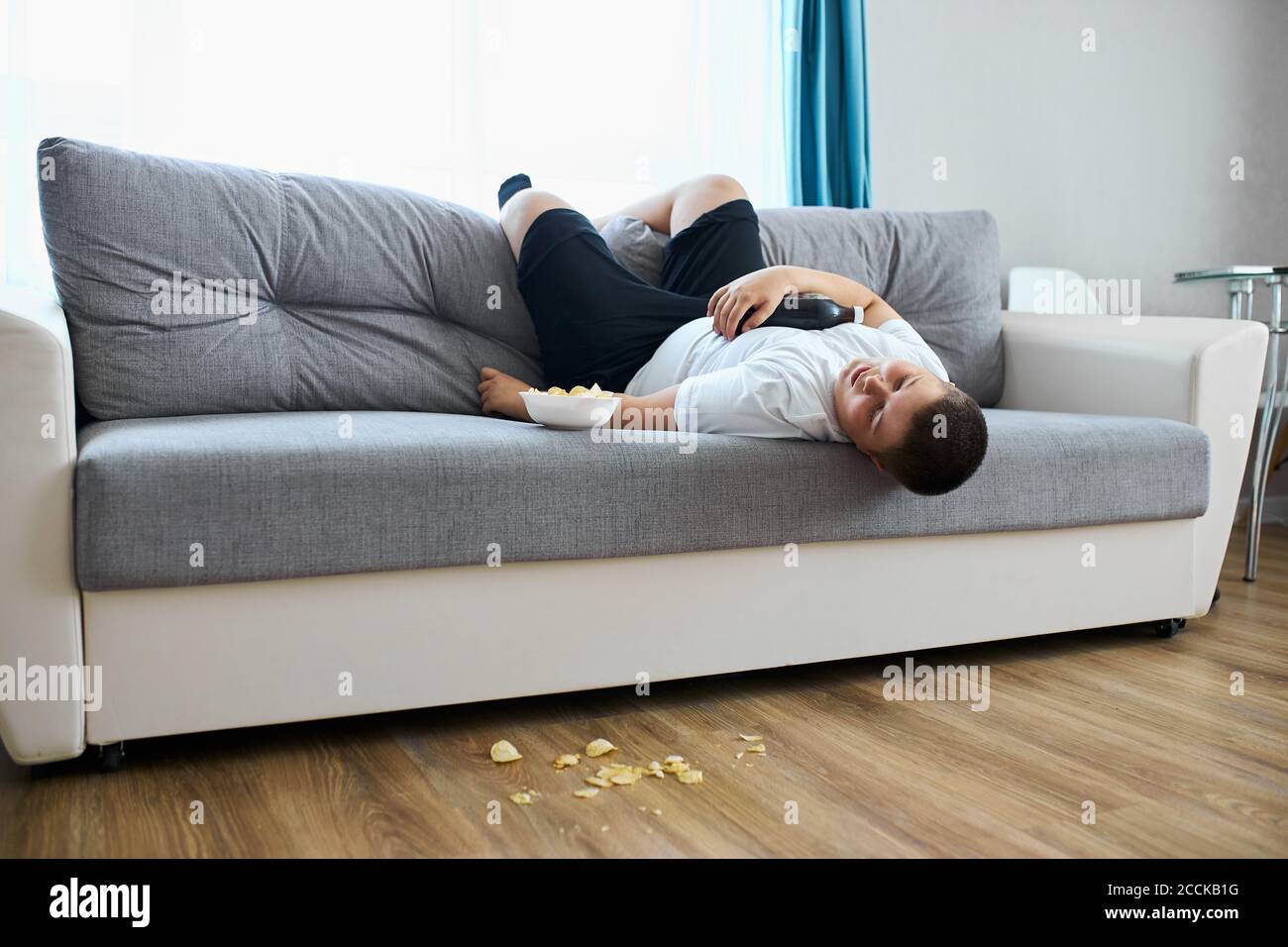 overeat fat boy sleep on sofa in living room, young caucasian teen boy fall asleep while he was watching tv and eating crisps Stock Photo