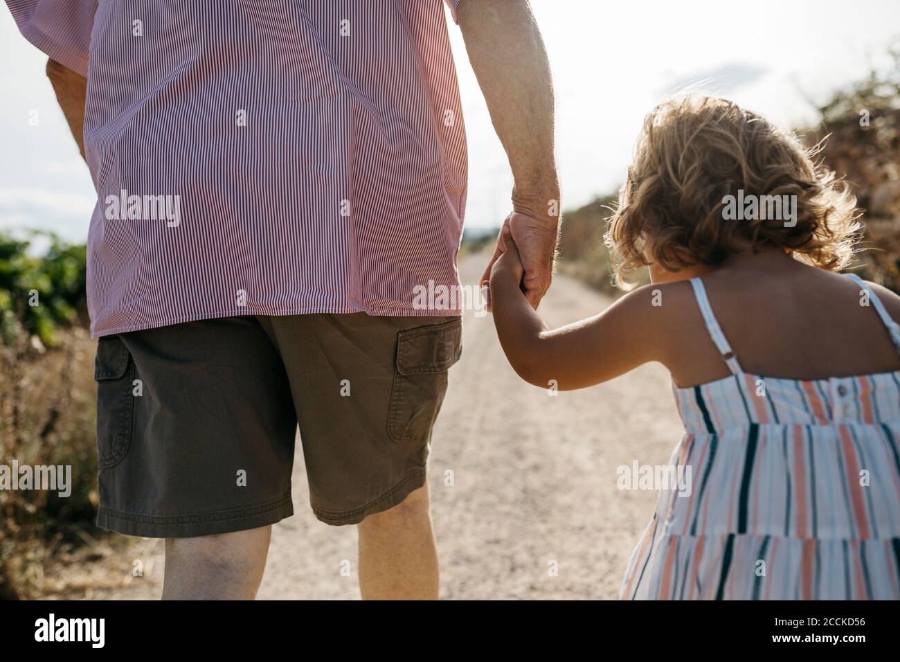 Close-up of grandfather holding granddaughter's hand while walking on dirt road Stock Photo