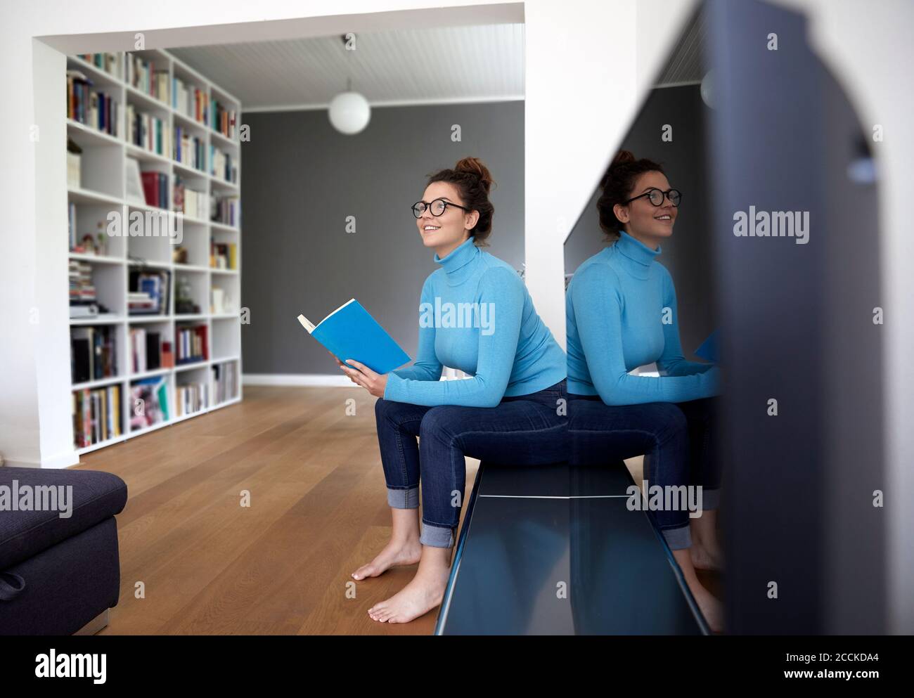 Smiling female student holding book and looking away while sitting by television set at home Stock Photo