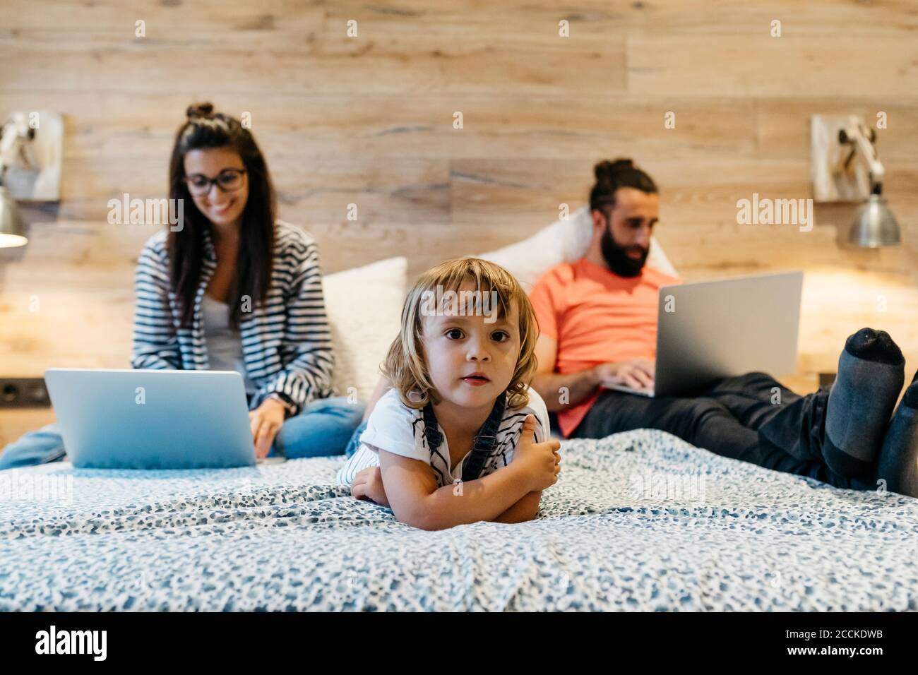 Cute girl lying on bed while mother working over laptop and father has fallen asleep in bedroom at home Stock Photo