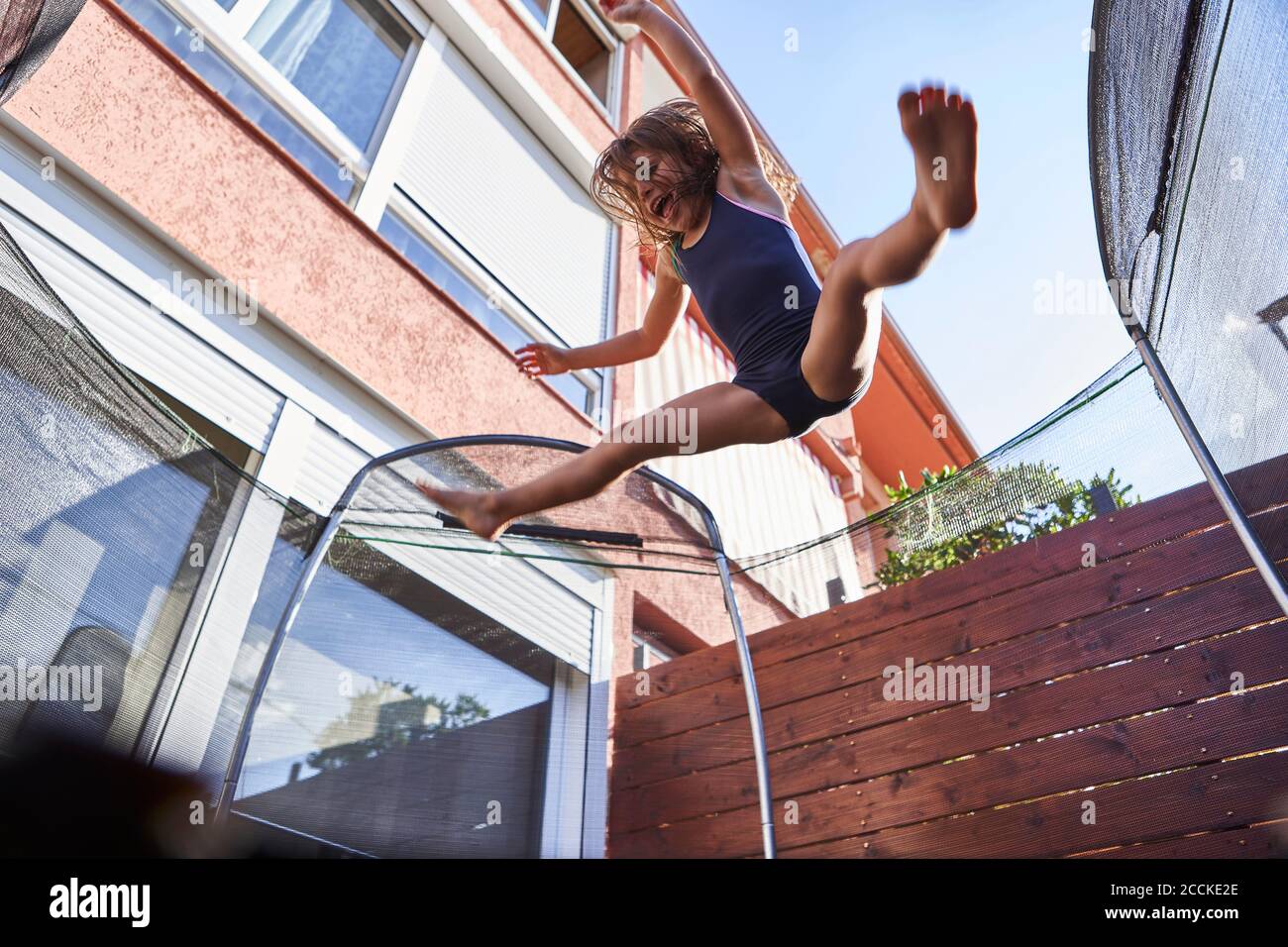 Cheerful girl jumping on trampoline against house during sunny day Stock Photo
