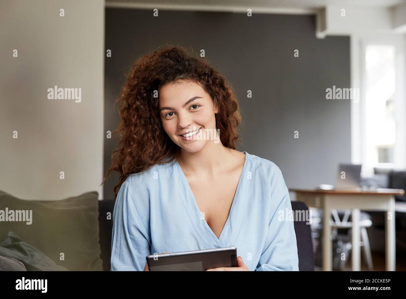 Smiling beautiful young woman with long brown curly hair holding digital tablet at home Stock Photo