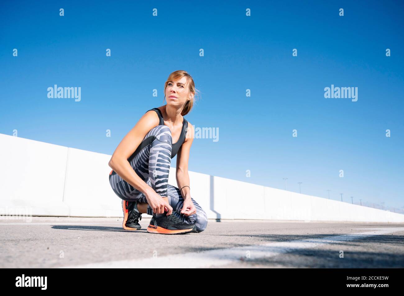 Mid adult woman looking away while tying shoelace on road against clear blue sky Stock Photo