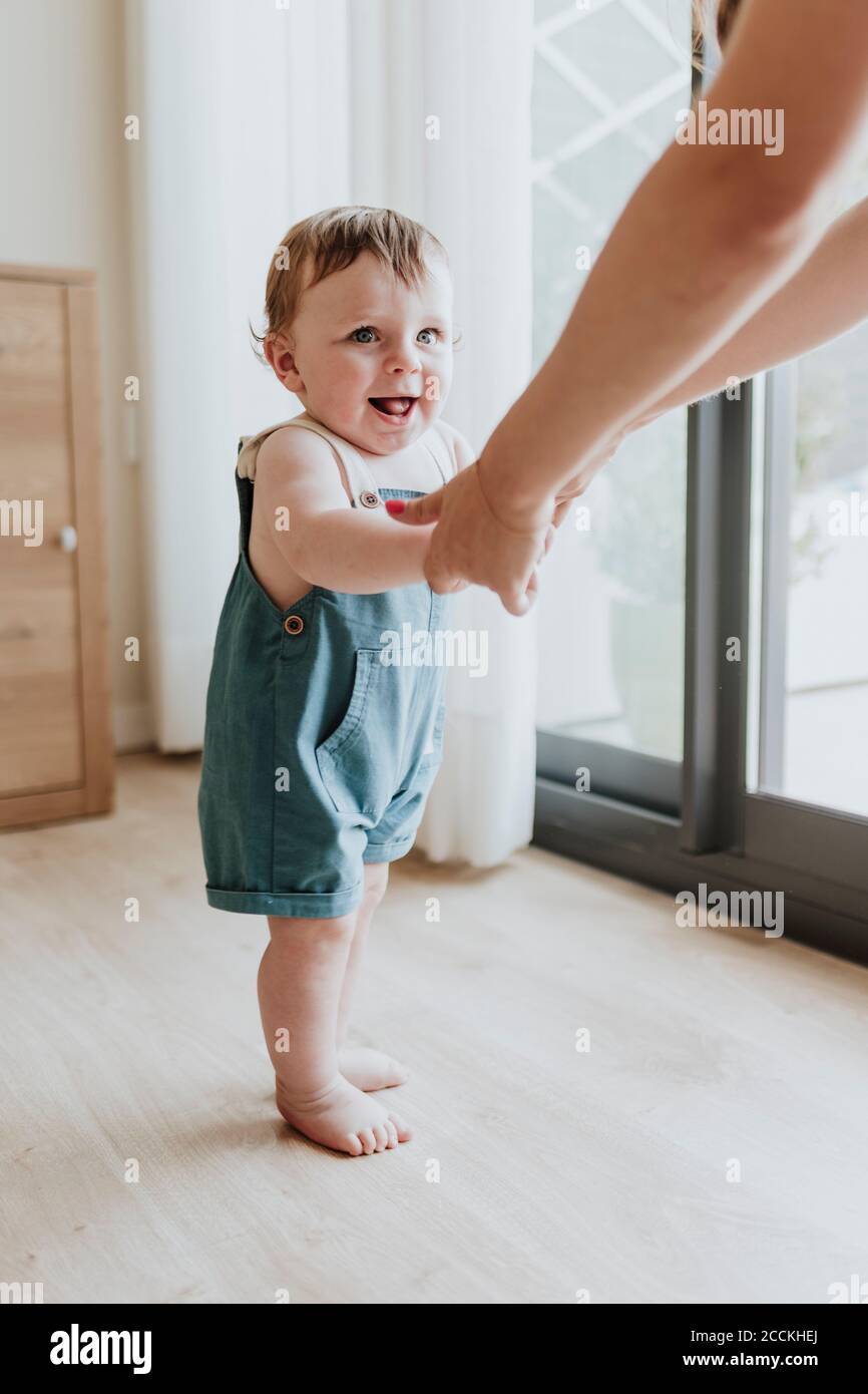 Baby girl holding mother's hands while learning to walk on floor at home Stock Photo